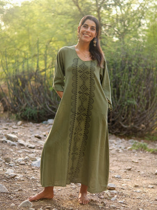 A woman stands barefoot on a rocky path outdoors, smiling, wearing the Garga Dress—a long olive green dress with geometric patterns—surrounded by green bushes and trees.