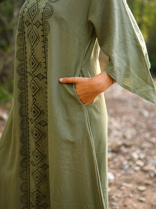 A person stands outdoors with one hand in the pocket, wearing the Garga Dress featuring black geometric embroidery on the front and loose sleeves, with a blurred rocky path and greenery in the background.