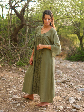 A woman stands barefoot on a rocky outdoor path, surrounded by trees, wearing the Garga Dress—a long, loose-fitting piece with detailed black embroidery. She gazes down and to the side amid natural greenery.