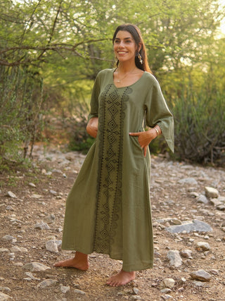Barefoot on a rocky path outdoors, a woman smiles in the olive-green Garga Dress with black geometric block prints. She has long dark hair, and lush green trees and sunlight surround her.