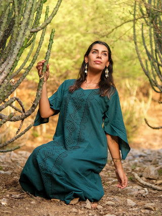 A woman wearing the Garga Dress with geometric prints kneels among tall cacti, gazing upward as sunlight filters through green foliage, creating a warm and serene atmosphere.