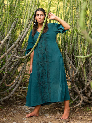 A woman in a teal geometric Garga Dress stands barefoot among tall green cacti, one arm raised as she gazes thoughtfully at the camera.