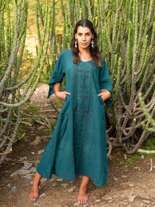 A woman with long dark hair stands barefoot on a rocky path among tall cacti, wearing the Garga Dress in teal with embroidered details and pockets, gazing directly at the camera.