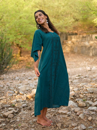A woman stands barefoot outdoors, smiling in a Garga Dress with teal embroidery and elbow-length sleeves. Green foliage and stone steps frame the rocky setting.