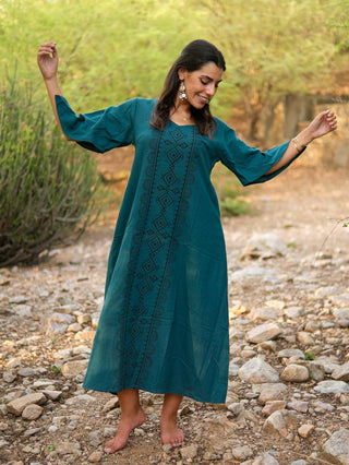 A woman smiles barefoot outdoors in a teal Garga Dress with geometric block prints, accessorized with earrings and a bracelet. She stands on rocky ground, arms outstretched, with greenery in the background.