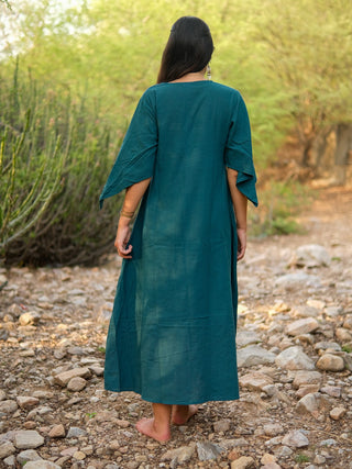 A woman with long dark hair wears the elegant teal Garga Dress, featuring wide sleeves and ankle length, as she stands barefoot on a rocky outdoor path surrounded by greenery, her back turned to the camera.