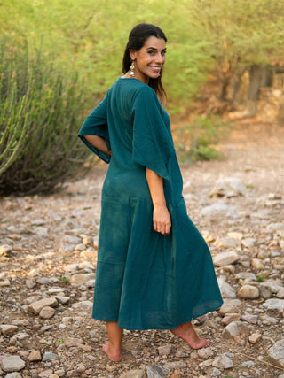 A woman with long dark hair, wearing the elegant Garga Dress and earrings, stands barefoot on a rocky outdoor path, smiling over her shoulder against a backdrop of green shrubs and trees.