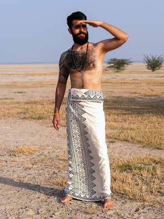 A man with a beard stands barefoot on dry grass and sand, wearing the Vyom Dhoti with intricate block prints. He shields his eyes while looking into the distance against a flat, open landscape under a clear sky.