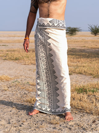 Standing on sandy ground, a person is wrapped in a Vyom Dhoti with black block print around the waist. Their upper body is bare, and their lower legs and feet are visible against a backdrop of dry landscape with sparse vegetation.