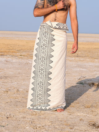 A person stands barefoot on sandy terrain wearing a Vyom Dhoti featuring intricate black geometric block prints. The traditional wrap-around garment leaves the chest exposed, complemented by a left arm tattoo and an orange wristband.