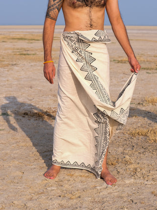 An individual stands barefoot on a sandy beach in a white Vyom Dhoti adorned with black geometric block prints. Their torso is bare, revealing a tattoo on one arm, set against a clear blue sky.