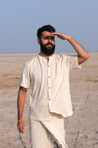 A bearded man wearing a natural-colored Indraja Shirt made from premium cotton and traditional attire stands outdoors on a dry landscape, shading his eyes with one hand as he gazes into the distance.