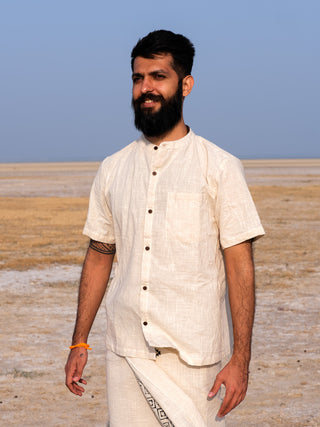 A bearded man in a Natural Indraja Shirt and traditional wrap stands outdoors on a dry, flat landscape under a clear blue sky, smiling softly.