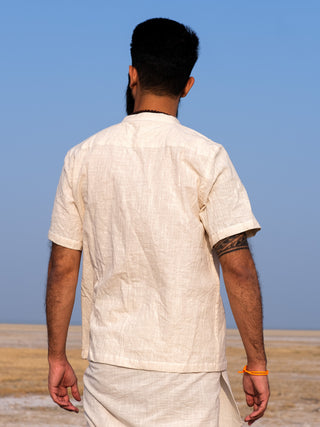 A man with short black hair and a beard, dressed in a premium cotton Indraja Shirt and traditional attire, stands outdoors facing away toward a flat, dry landscape beneath a clear blue sky.