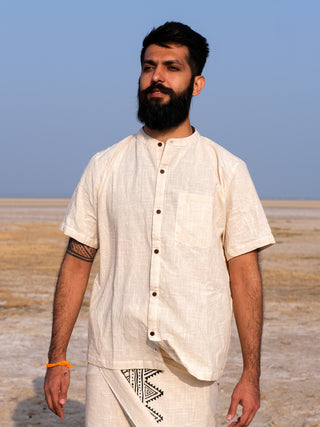 A bearded man with dark hair stands outdoors on a dry, flat landscape under a clear blue sky. He wears the Indraja Shirt with matching geometric-patterned pants, made from premium cotton, plus a visible tattoo and orange bracelet.