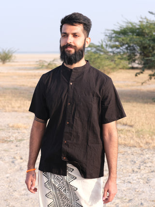 A man with a dark beard and short hair stands outdoors on a sandy, grassy area, wearing the black Indraja Shirt and white pants with a black geometric pattern. Trees and an open landscape are seen in the background.
