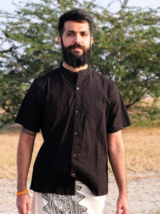 A bearded man wears the black Indraja Shirt and a patterned white wrap, standing outdoors with dry grass and green trees behind him.