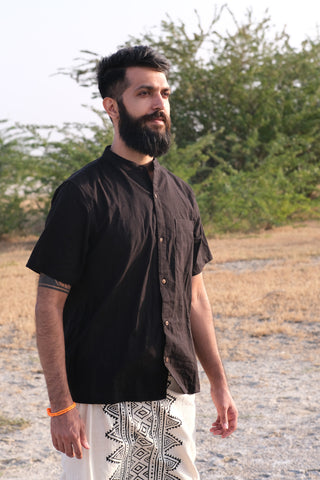 A bearded man wearing a premium quality black Indraja Shirt and patterned white pants stands outdoors on dry grass, with green bushes and a pale sky in the background.