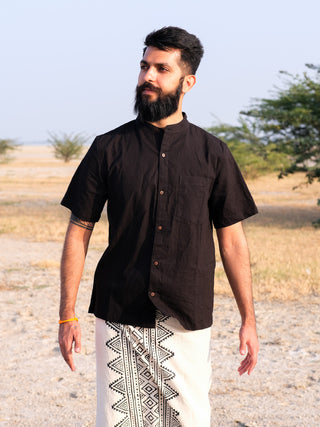 A bearded man stands outside on dry grass, wearing an Indraja Shirt—black with white geometric patterns, short-sleeved and made from premium cotton. Trees and an open field appear in the background.