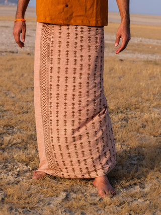 A person stands barefoot on dry grass, wearing the Rama Dhoti—a peach-colored block print traditional garment—and an orange shirt, with only the lower body visible.
