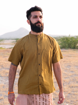 A bearded man wearing a green Indraja Shirt made from premium cotton and traditional patterned pants stands outdoors on dry grass with hills and greenery in the background, gazing confidently into the distance.