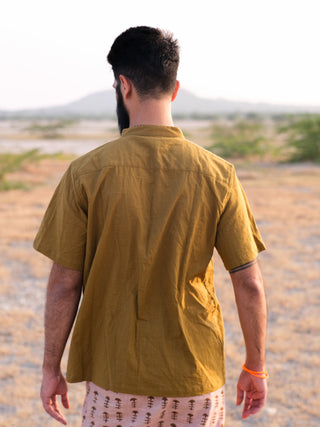 A man with short dark hair and a beard wears the Indraja Shirt in mustard yellow, standing outdoors with dry grass, bushes, and hills in the background.