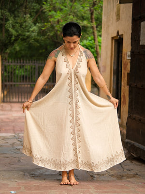 A woman stands barefoot outside among trees and a nearby building, holding out the sides of her beige Hanvi maxi dress. Sleeveless and adorned with a delicate block print and patterned trim, the dress is crafted from sustainable cotton, perfectly complementing the natural setting.