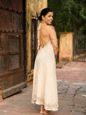 A woman, barefoot on a stone path and smiling over her shoulder, wears the Hanvi maxi dress—a cream, backless design crafted from sustainable cotton with delicate block print details. The dress accentuates her tattoos and dark hair tied back, while a rustic wooden door and lush greenery provide an inviting backdrop.