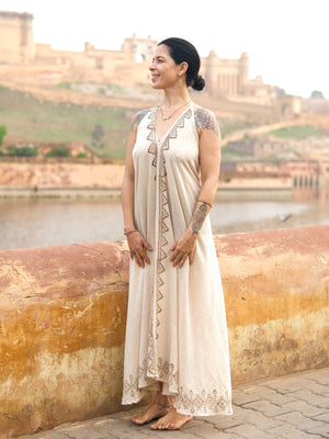 Wearing the Hanvi maxi dress—a sleeveless piece crafted from sustainable cotton and adorned with block print geometric patterns—a woman stands by a river, smiling. Her tattoos are visible on her arms as the dress beautifully complements the old buildings and green hills in the background.