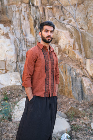 A man with a beard, wearing the rust-colored Acharya Shirt Full-sleeve and black pants, stands with hands in pockets against a rocky cliff. His expression is thoughtful as he gazes into the distance.