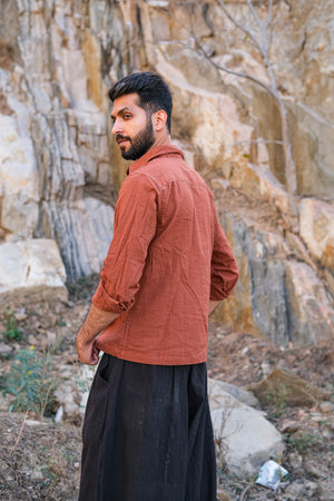 A bearded man stands outdoors against a rocky backdrop, wearing the Acharya Shirt Full-sleeve made from organic cotton and dark pants. He glances back, his hands by his sides, amid sparse vegetation.