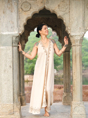 A woman in a long, natural Hanvi maxi dress with tattoos on her arms stands barefoot in an ornate stone archway, smiling and looking to the side. Crafted from sustainable cotton with a delicate block print, her dress complements the greenery and water visible in the background.
