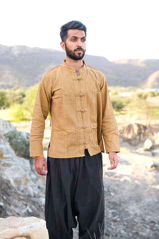 A man with short hair and a beard stands on rocky terrain outdoors, wearing a boho-inspired Knight Shirt and loose black pants, with hills and greenery in the background.