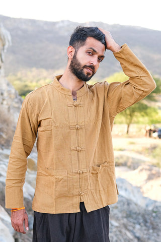 A bearded man outdoors wears the tan Knight Shirt made from sustainable cotton and dark pants, touching his head with one hand. Rocky terrain and distant hills appear in the background.