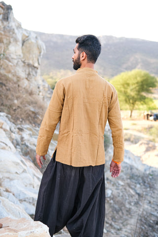 A man stands on rocky terrain, facing away from the camera. He wears a tan Knight Shirt made from sustainable cotton and loose black pants, with hills, rocks, and trees in the background.