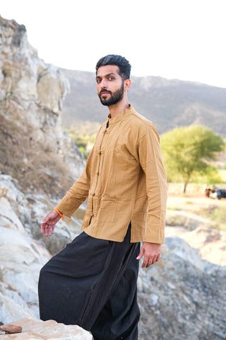 A bearded man, dressed in a tan Knight Shirt crafted from sustainable cotton and loose black pants, stands outdoors on rocky terrain with hills and trees behind him.
