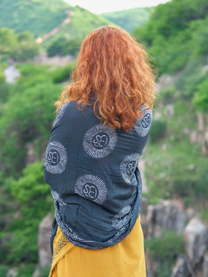 A person with long curly red hair stands outdoors facing green hills and rocky terrain, draped in the luxurious Mantra Shawl featuring white sun patterns.
