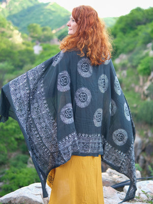 A woman with long red hair, wearing a yellow dress and a luxurious black Mantra Shawl with white patterns, stands outdoors on rocks with a green, mountainous background.
