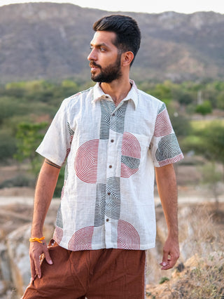 A stylish man with a beard stands outdoors among hills and greenery, looking to his left. He wears the white Darlion Shirt with circular block prints and brown pants.