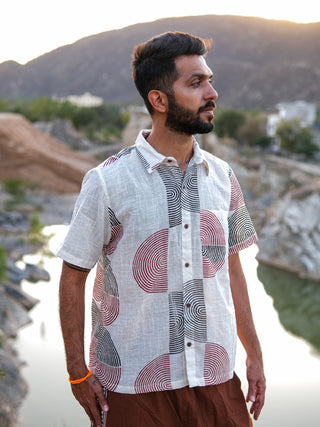 A bearded man stands outdoors by a river and mountains at sunset, looking to the side in a short-sleeve Darlion Shirt with circular block prints and brown pants.