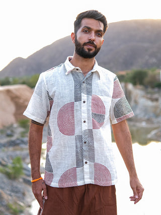 A man with short dark hair and a beard stands outdoors near rocky hills and water, wearing the stylish Darlion Shirt with red and black circular block-printed patterns, paired with brown pants.