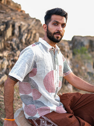A bearded man sits on a rock outdoors, wearing the stylish Darlion Shirt with red and black circular block print patterns and brown pants. Rocky terrain and greenery are in the background.