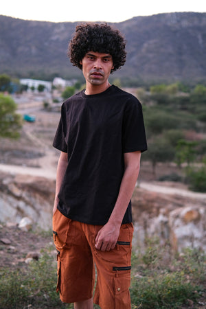 A person with curly hair wearing a stylish black Alec T-shirt and orange cargo shorts stands outdoors in a natural, rocky landscape with hills and greenery in the background.
