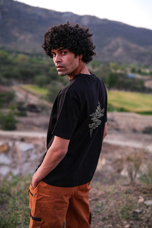 A man with curly hair stands outdoors, looking over his shoulder. He wears a stylish black Alec T-shirt with a floral graphic on the back and brown pants. Mountains and greenery are visible in the background.