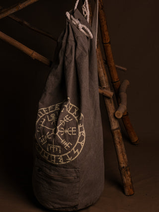 The Vegvisir Duffle bag is a gray drawstring bag featuring white Norse runes and a circular Vegvisir symbol, providing secure storage for travel. It’s pictured leaning against bamboo poles on a dark brown background.