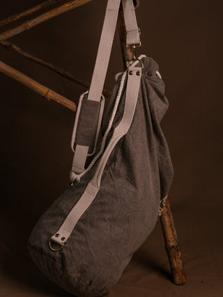 The Vegvisir Duffle bag, featuring a gray design and white straps, hangs from a wooden structure against a brown background—perfect for secure storage and ideal as your traveling companion.