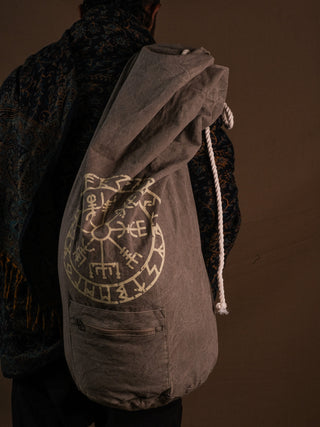 A person in a patterned shawl carries the grey Vegvisir Duffle bag, which features a white circular rune symbol and provides secure storage, set against a plain brown background.