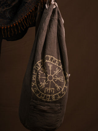 The Vegvisir Duffle bag, in large gray with a circular symbol and runic design, hangs against a brown background. A patterned textile appears at the top left. This secure and unique travel companion is perfect for your next trip.