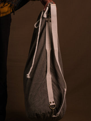 A person holds the Vegvisir Duffle bag—large and gray with white straps and a rope handle—against a brown background, perfect for secure storage on any adventure. Only their hand, torso, and leg are visible.