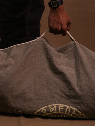 A hand and lower arm, wearing a black watch and dark pants, hold the large gray Vegvisir Duffle bag by its white rope handle against a brown backdrop.